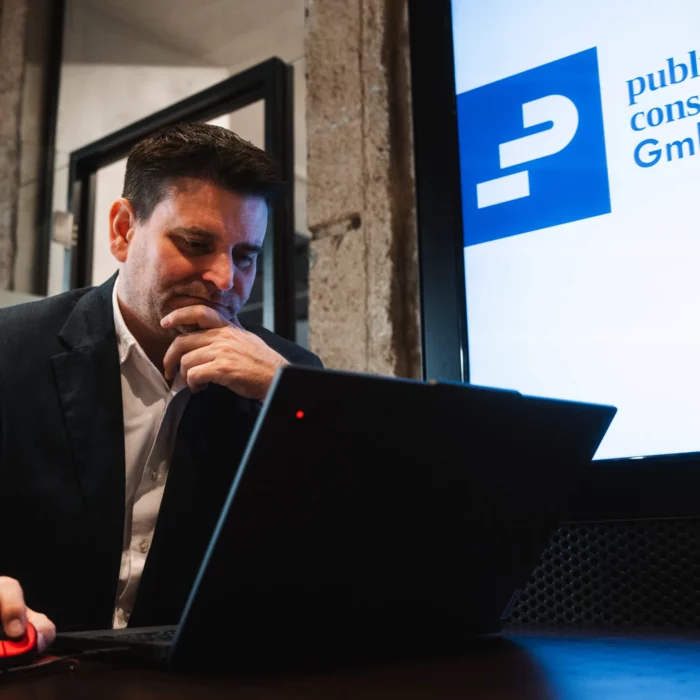 Nachdenkliches Teammitglied von public.consulting am Laptop vor Bildschirm mit Firmenlogo