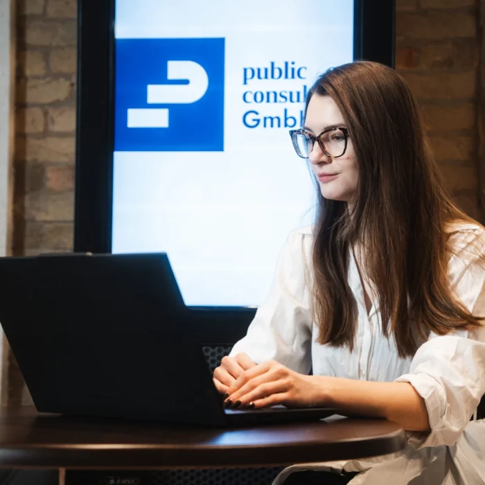 Teammitglied von public.consulting arbeitet am Laptop vor einem Bildschirm mit Firmenlogo