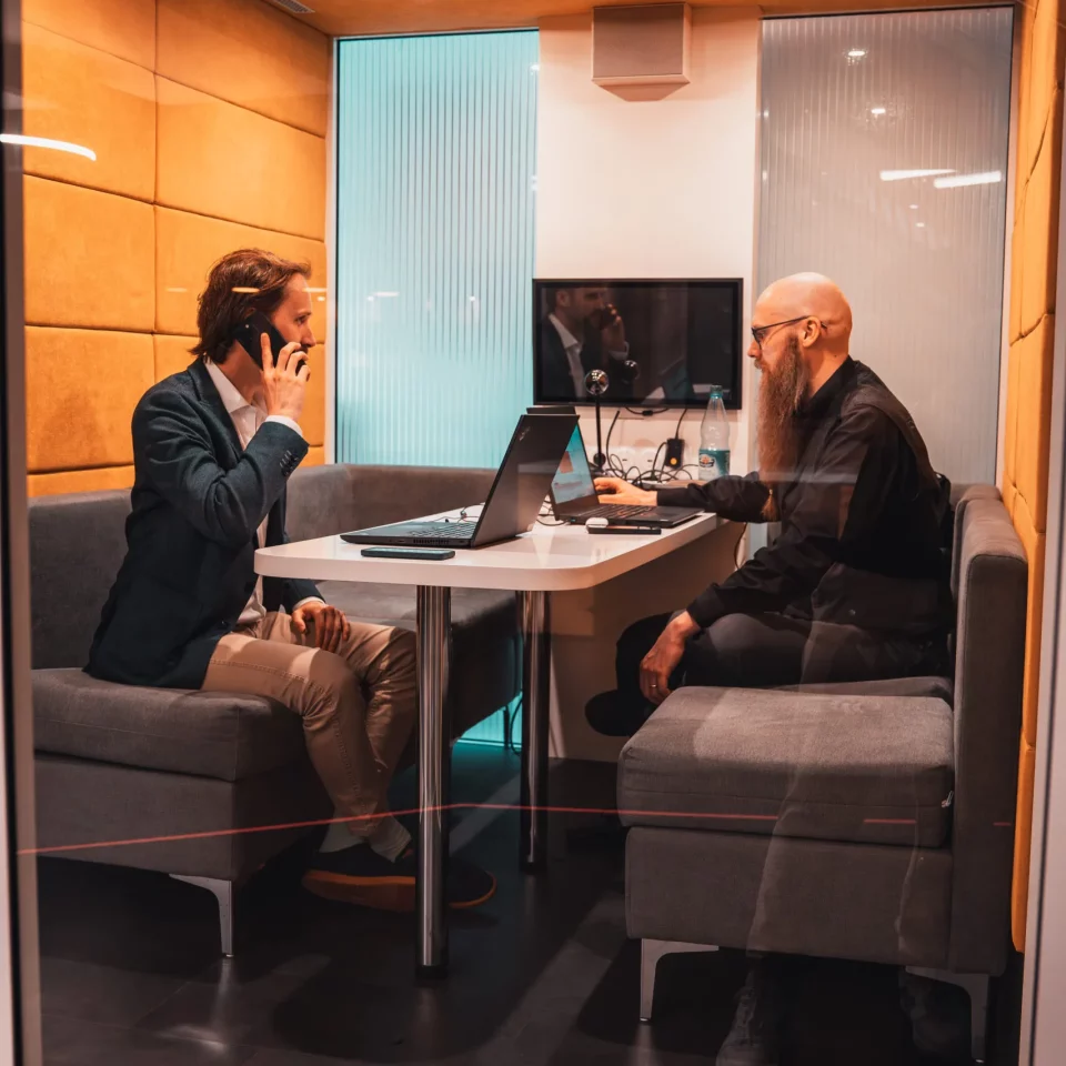 Zwei Teammitglieder von public.consulting arbeiten in einer Telefonbox mit Laptops